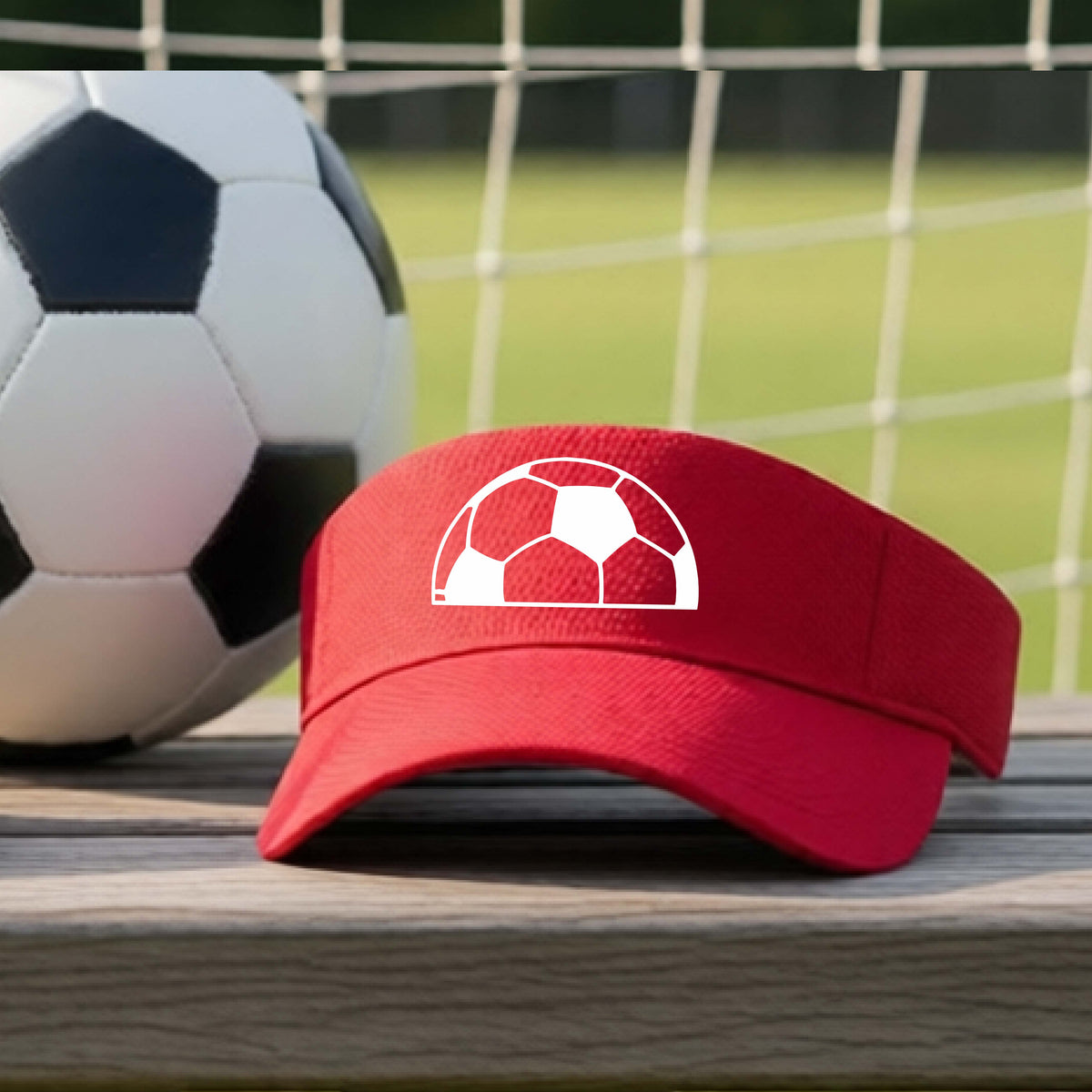A red baseball cap with a white soccer ball graphic on the front, resting on a wooden surface with a soccer ball and a goal net in the blurred background.