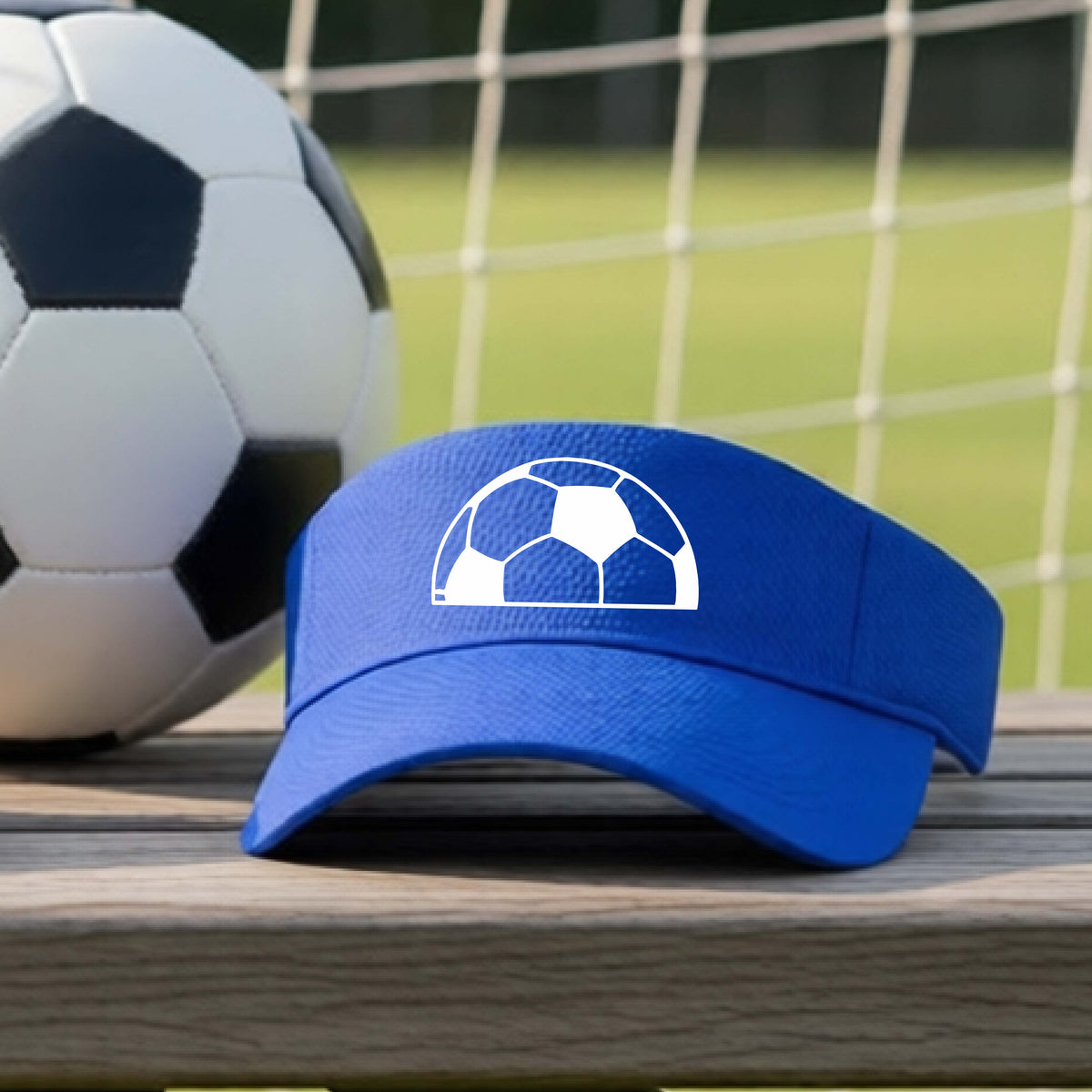 A royal blue baseball cap with a white soccer ball graphic on the front, resting on a wooden surface with a soccer ball and a goal net in the blurred background.