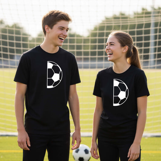 Two young people standing on a soccer field wearing matching black t-shirts with a white half soccer ball graphic, with a soccer net and grass in the background.