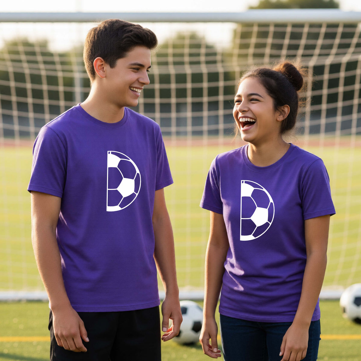Two young people standing on a soccer field wearing matching purple t-shirts with a white half soccer ball graphic, with a soccer net and grass in the background.