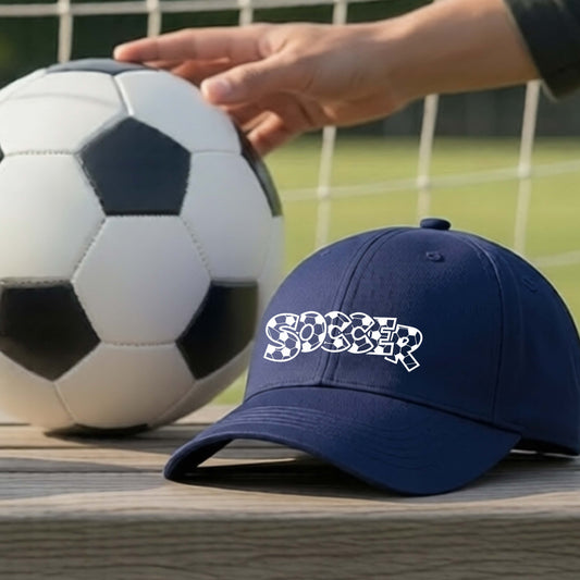 A close-up of a navy blue hat sitting on an outdoor bench next to a soccer ball in front of goal, with a white graphic of the word 'SOCCER' in a stylish soccer pattern printed on the front.