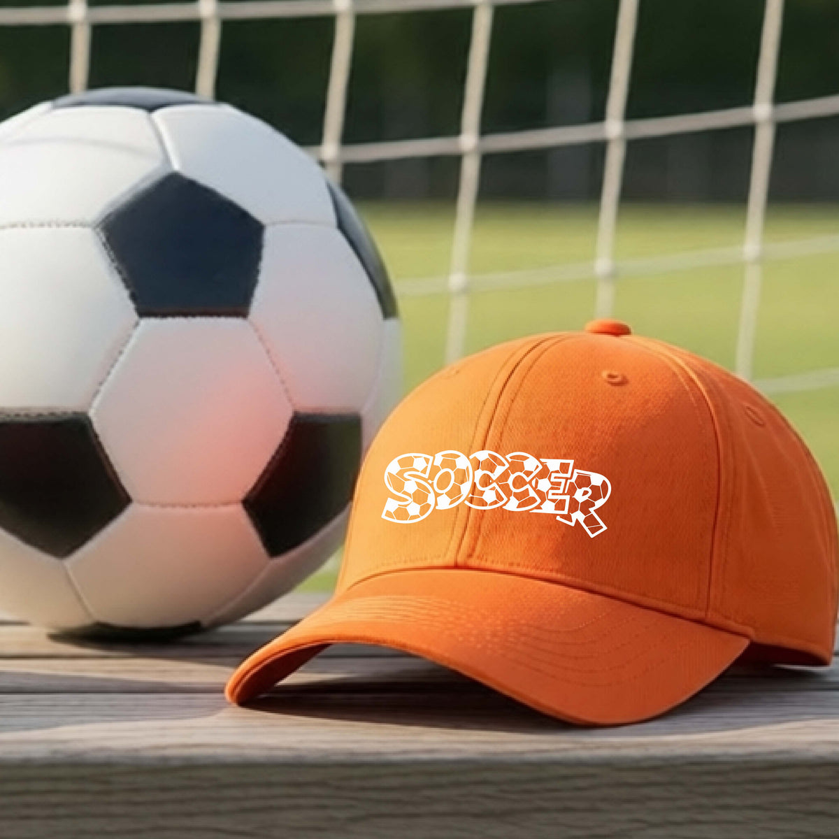 A close-up of an orange hat sitting on an outdoor bench next to a soccer ball in front of goal, with a white graphic of the word 'SOCCER' in a stylish soccer pattern printed on the front.