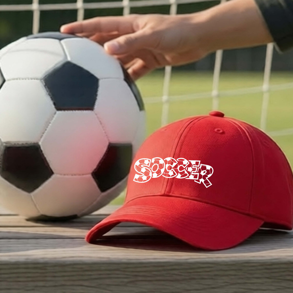 A close-up of a red hat sitting on an outdoor bench next to a soccer ball in front of goal, with a white graphic of the word 'SOCCER' in a stylish soccer pattern printed on the front.
