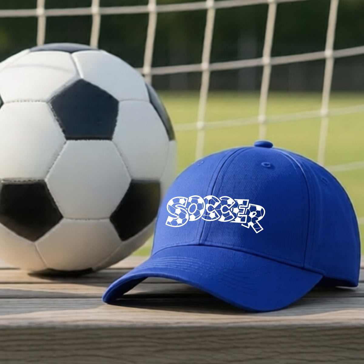 A close-up of a royal blue hat sitting on an outdoor bench next to a soccer ball in front of goal, with a white graphic of the word 'SOCCER' in a stylish soccer pattern printed on the front.