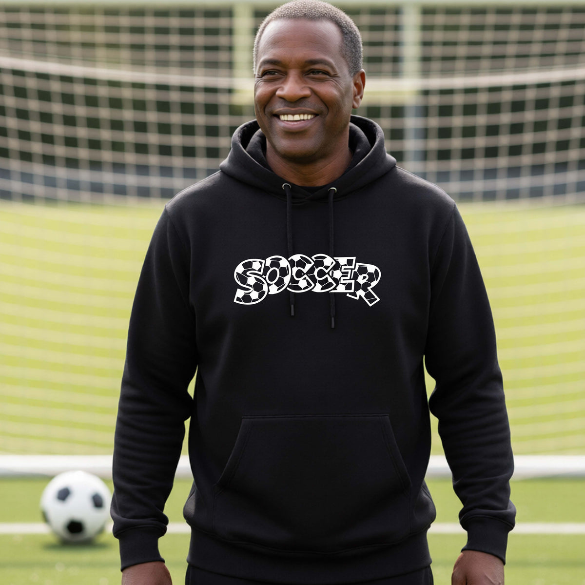 A smiling man wearing a black hoodie pullover , standing on a soccer field in front of a goal net. with a white graphic of the word 'SOCCER' in a stylish soccer pattern printed on the chest.
