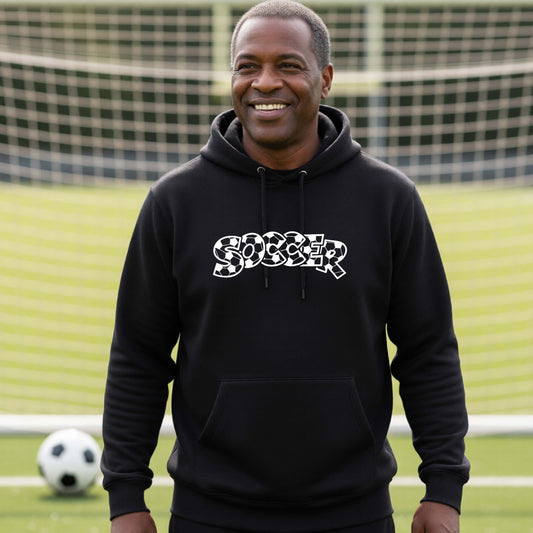 A smiling man wearing a black hoodie pullover , standing on a soccer field in front of a goal net. with a white graphic of the word 'SOCCER' in a stylish soccer pattern printed on the chest.