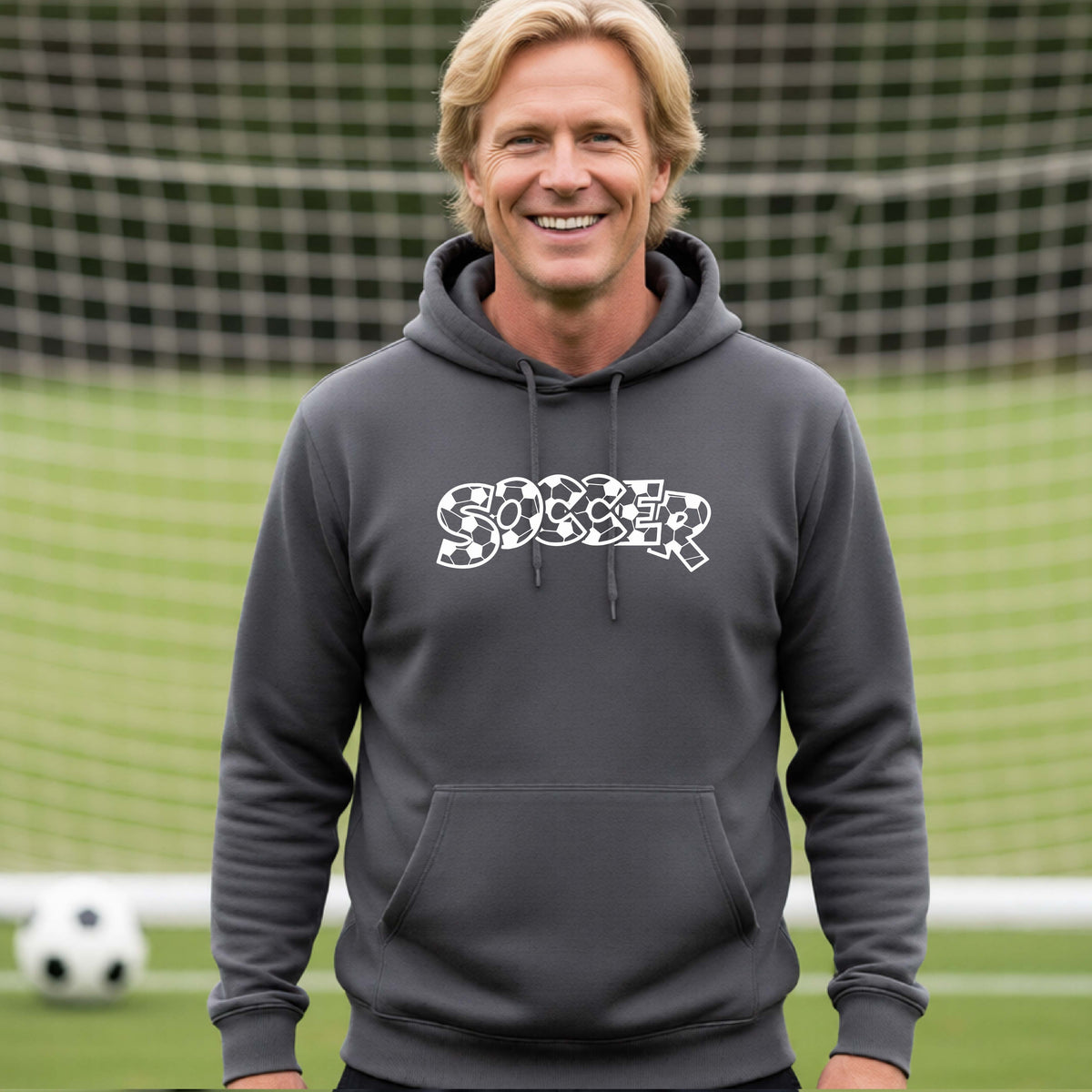 A smiling man wearing a gray hoodie pullover , standing on a soccer field in front of a goal net. with a white graphic of the word 'SOCCER' in a stylish soccer pattern printed on the chest.