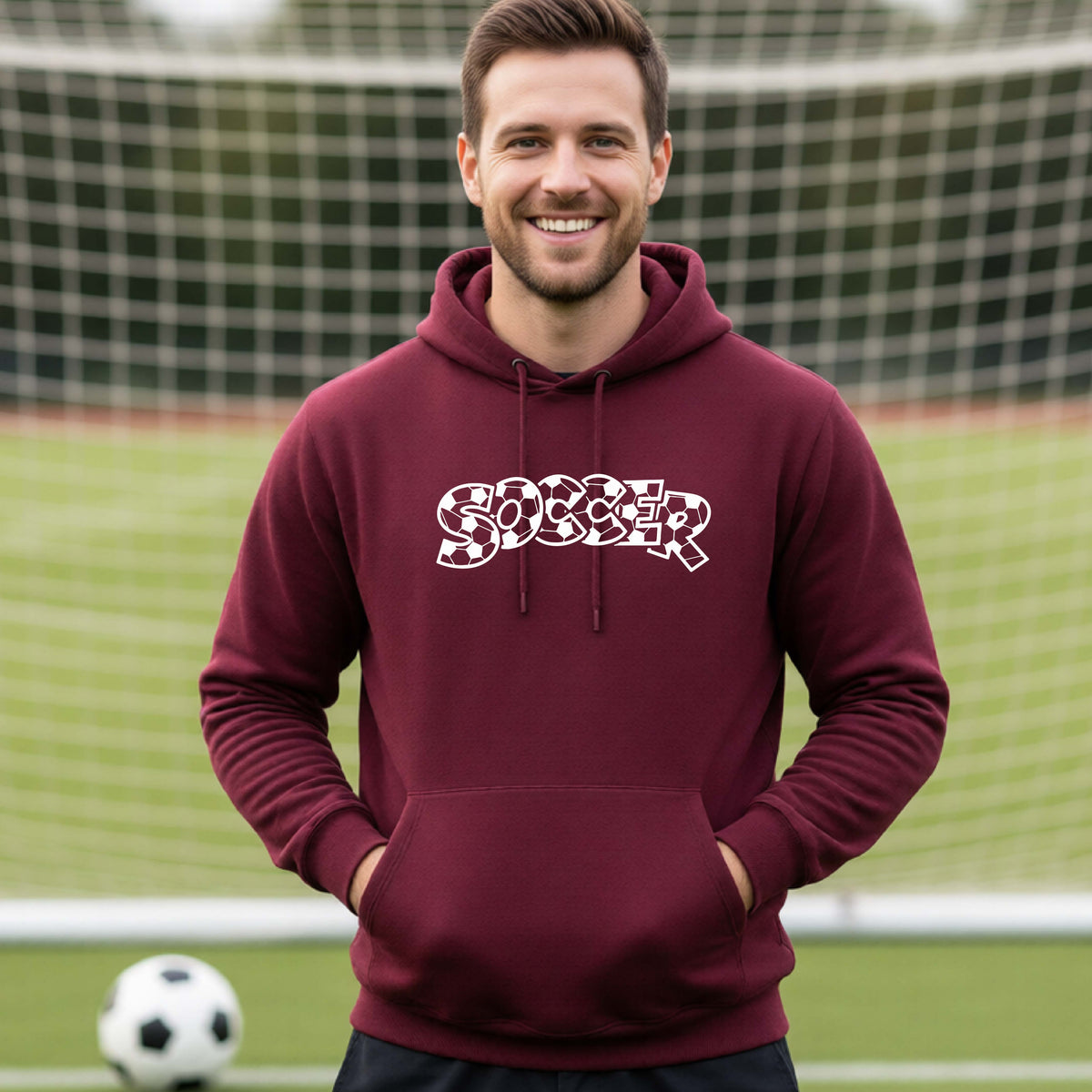 A smiling man wearing a maroon hoodie pullover , standing on a soccer field in front of a goal net. with a white graphic of the word 'SOCCER' in a stylish soccer pattern printed on the chest.