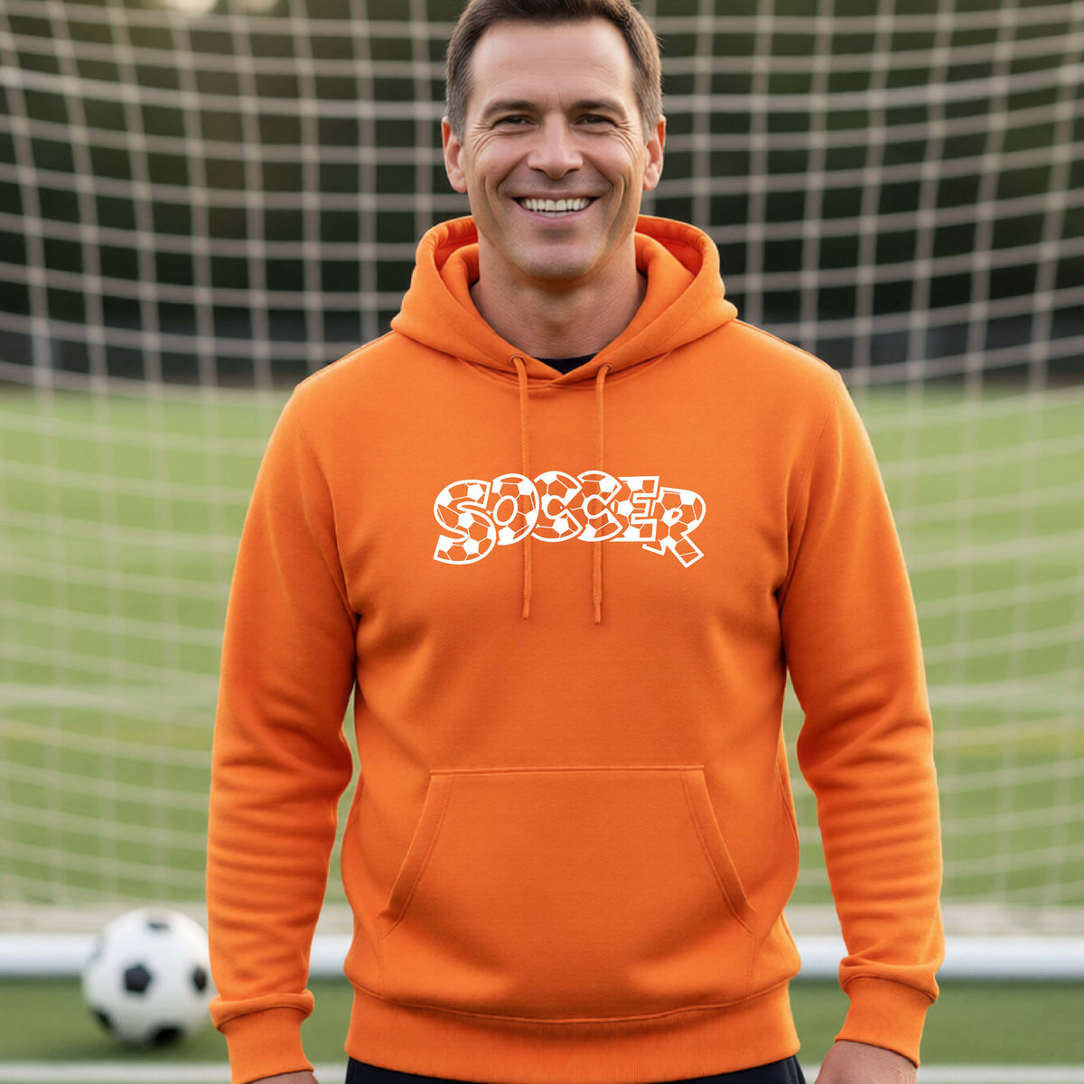 A smiling man wearing an orange hoodie pullover , standing on a soccer field in front of a goal net. with a white graphic of the word 'SOCCER' in a stylish soccer pattern printed on the chest.