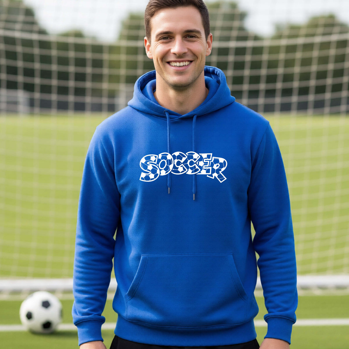 A smiling man wearing a royal blue hoodie pullover , standing on a soccer field in front of a goal net. with a white graphic of the word 'SOCCER' in a stylish soccer pattern printed on the chest.