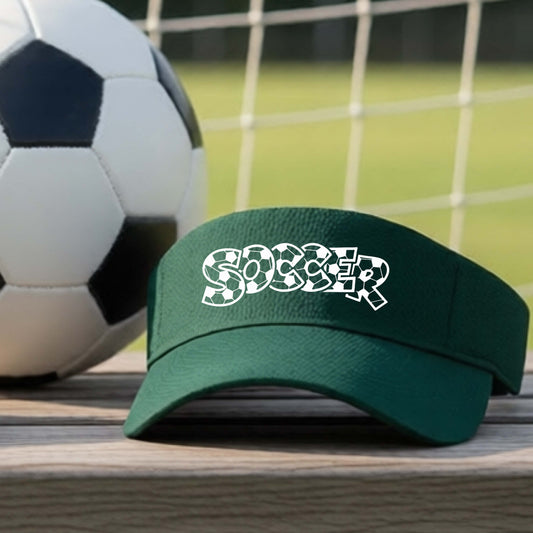 A close-up of a green visor sitting on an outdoor bench next to a soccer ball in front of goal, with a white graphic of the word 'SOCCER' in a stylish soccer pattern printed on the front. 