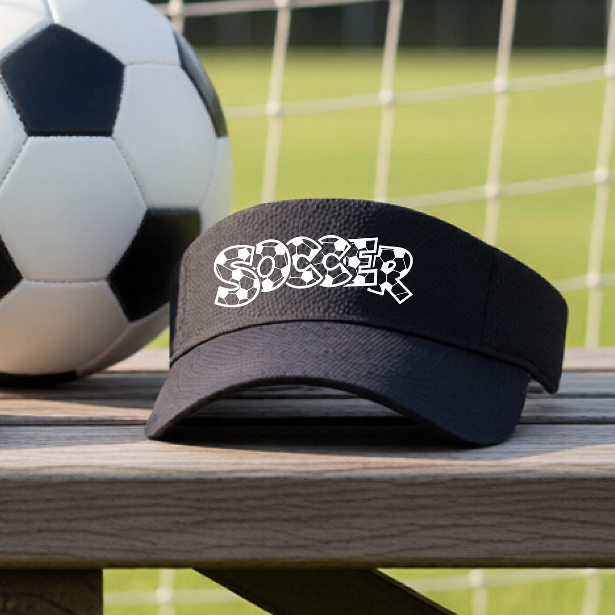 A close-up of a  black visor sitting on an outdoor bench next to a soccer ball in front of goal, with a white graphic of the word 'SOCCER' in a stylish soccer pattern printed on the front. 