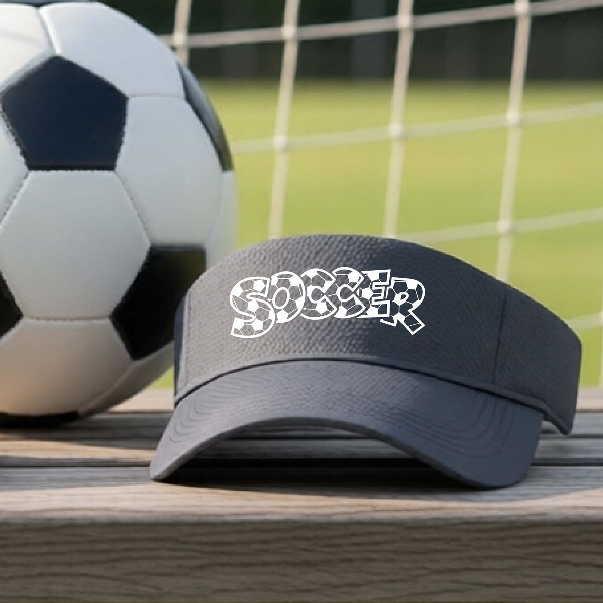 A close-up of a gray visor sitting on an outdoor bench next to a soccer ball in front of goal, with a white graphic of the word 'SOCCER' in a stylish soccer pattern printed on the front. 