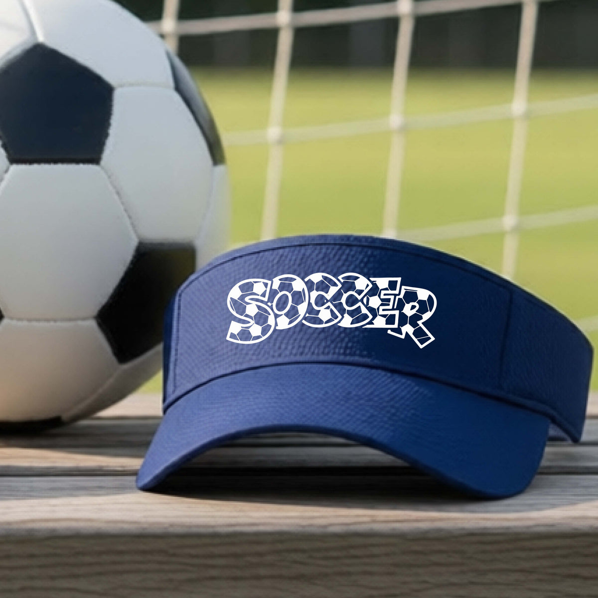A close-up of a navy blue visor sitting on an outdoor bench next to a soccer ball in front of goal, with a white graphic of the word 'SOCCER' in a stylish soccer pattern printed on the front. 