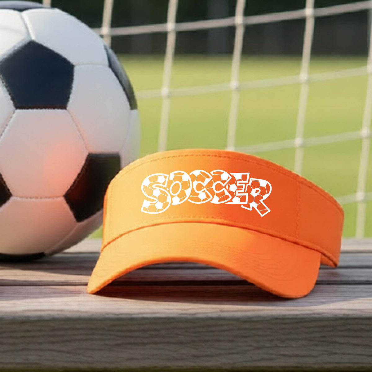 A close-up of an orange visor sitting on an outdoor bench next to a soccer ball in front of goal, with a white graphic of the word 'SOCCER' in a stylish soccer pattern printed on the front. 