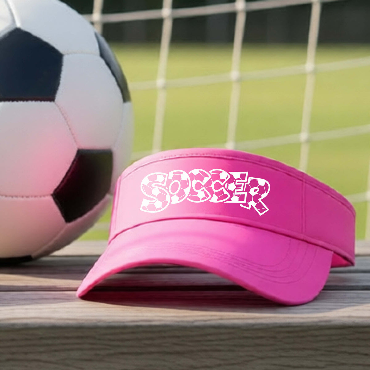 A close-up of a pink visor sitting on an outdoor bench next to a soccer ball in front of goal, with a white graphic of the word 'SOCCER' in a stylish soccer pattern printed on the front. 