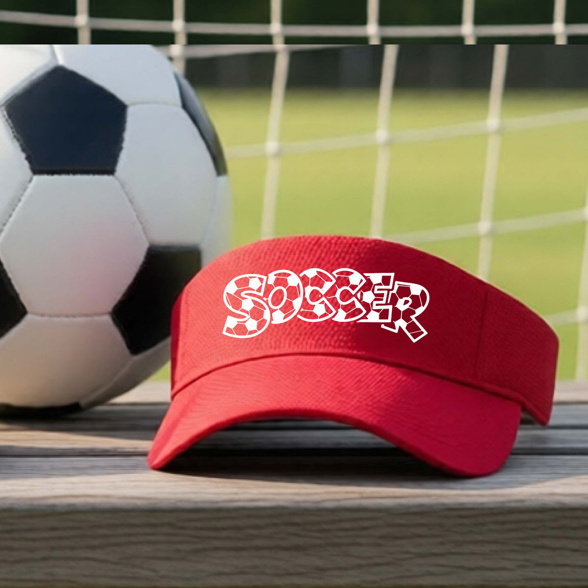 A close-up of a red visor sitting on an outdoor bench next to a soccer ball in front of goal, with a white graphic of the word 'SOCCER' in a stylish soccer pattern printed on the front. 