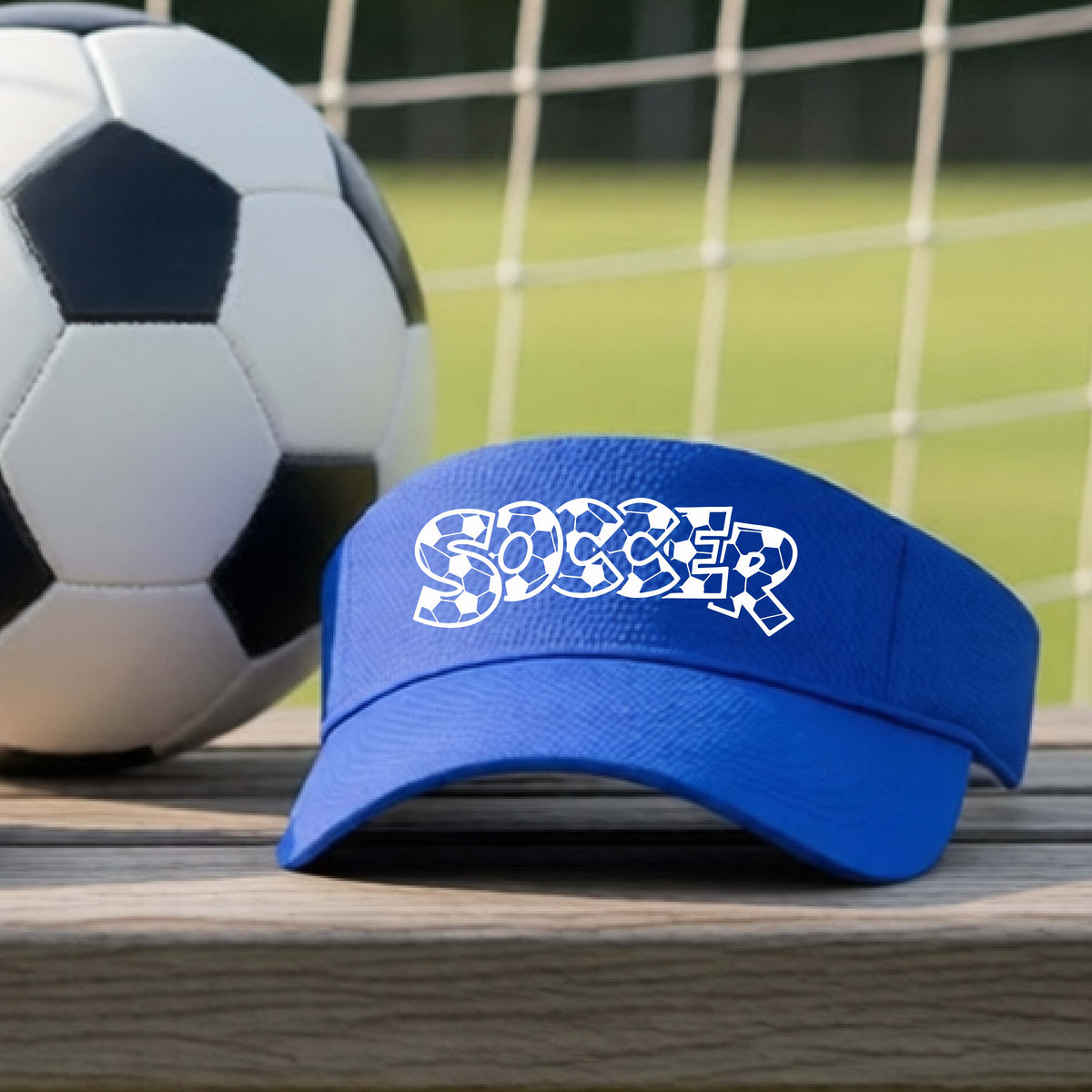 A close-up of a royal blue visor sitting on an outdoor bench next to a soccer ball in front of goal, with a white graphic of the word 'SOCCER' in a stylish soccer pattern printed on the front. 