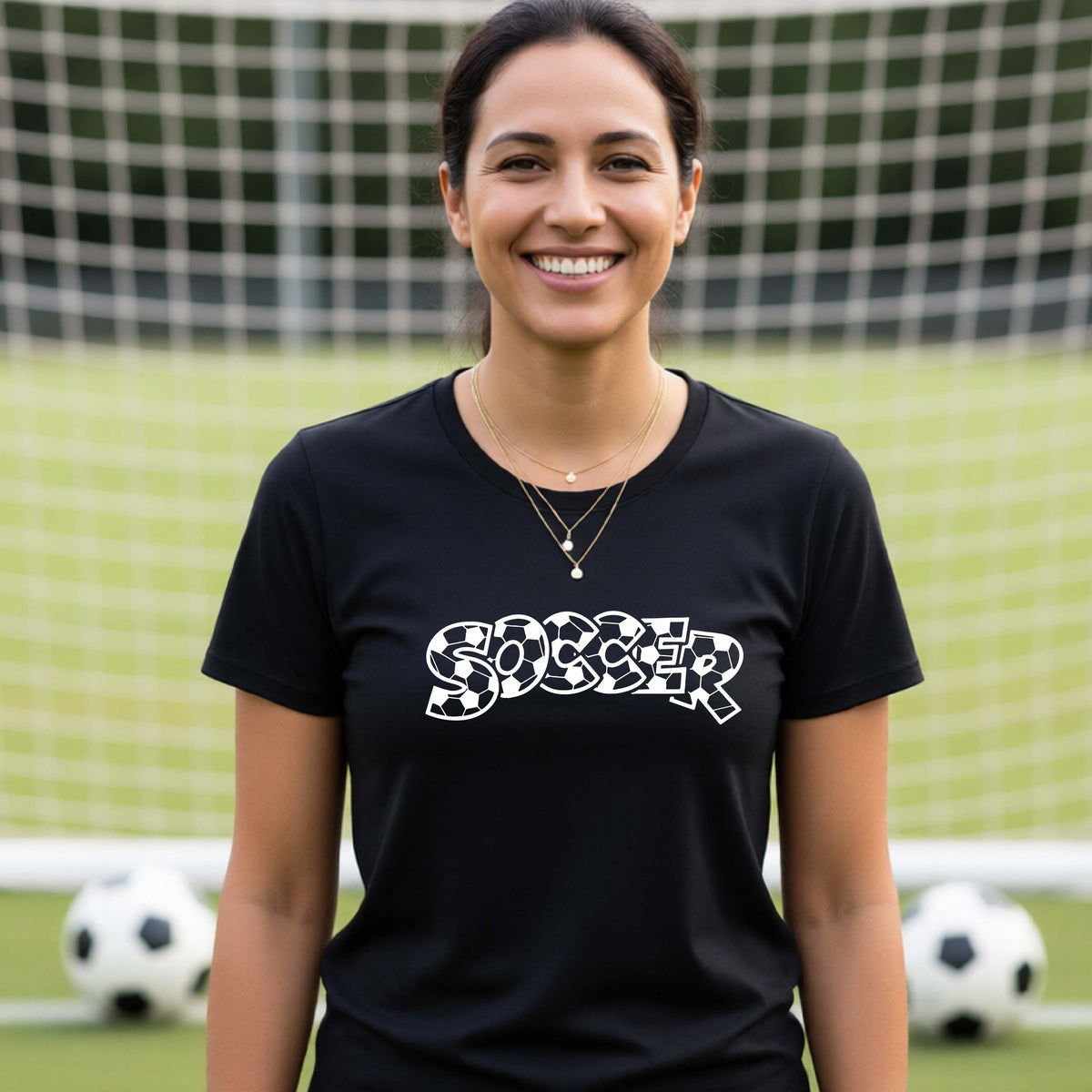 A smiling woman wearing a black t-shirt, standing on a soccer field in front of a goal net. with a white graphic of the word 'SOCCER' in a stylish soccer pattern printed on the chest.