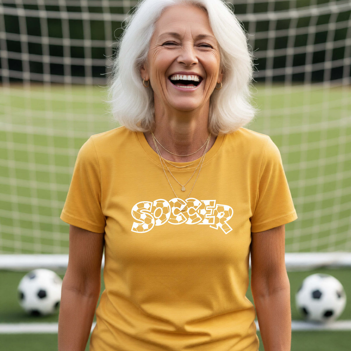 A smiling woman wearing a gold t-shirt, standing on a soccer field in front of a goal net. with a white graphic of the word 'SOCCER' in a stylish soccer pattern printed on the chest.