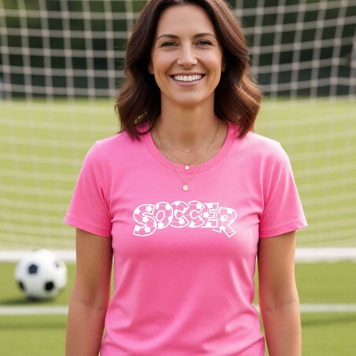 A smiling woman wearing a pink t-shirt, standing on a soccer field in front of a goal net. with a white graphic of the word 'SOCCER' in a stylish soccer pattern printed on the chest.