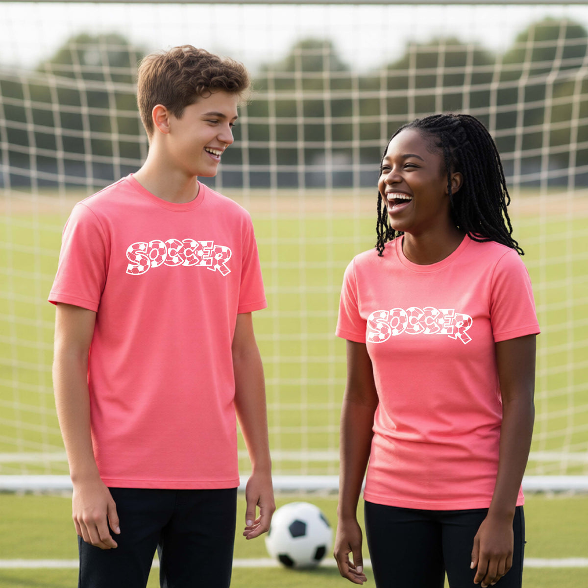 Two young athletics wearing matching coral pink t-shirt, standing on a soccer field in front of a goal net. with a white graphic of the word 'SOCCER' in a stylish soccer pattern printed on the chests.