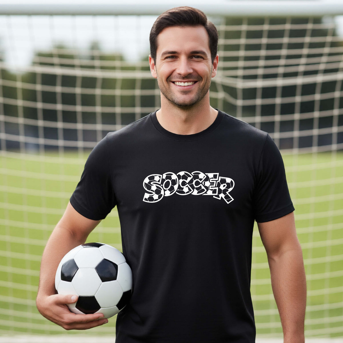 A smiling man wearing a black t-shirt, standing on a soccer field in front of a goal net. with a white graphic of the word 'SOCCER' in a stylish soccer pattern printed on the chest, holding a soccer ball.