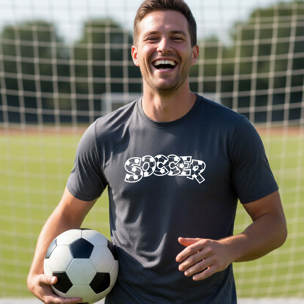 A smiling man wearing a dark gray t-shirt, standing on a soccer field in front of a goal net. with a white graphic of the word 'SOCCER' in a stylish soccer pattern printed on the chest, holding a soccer ball.