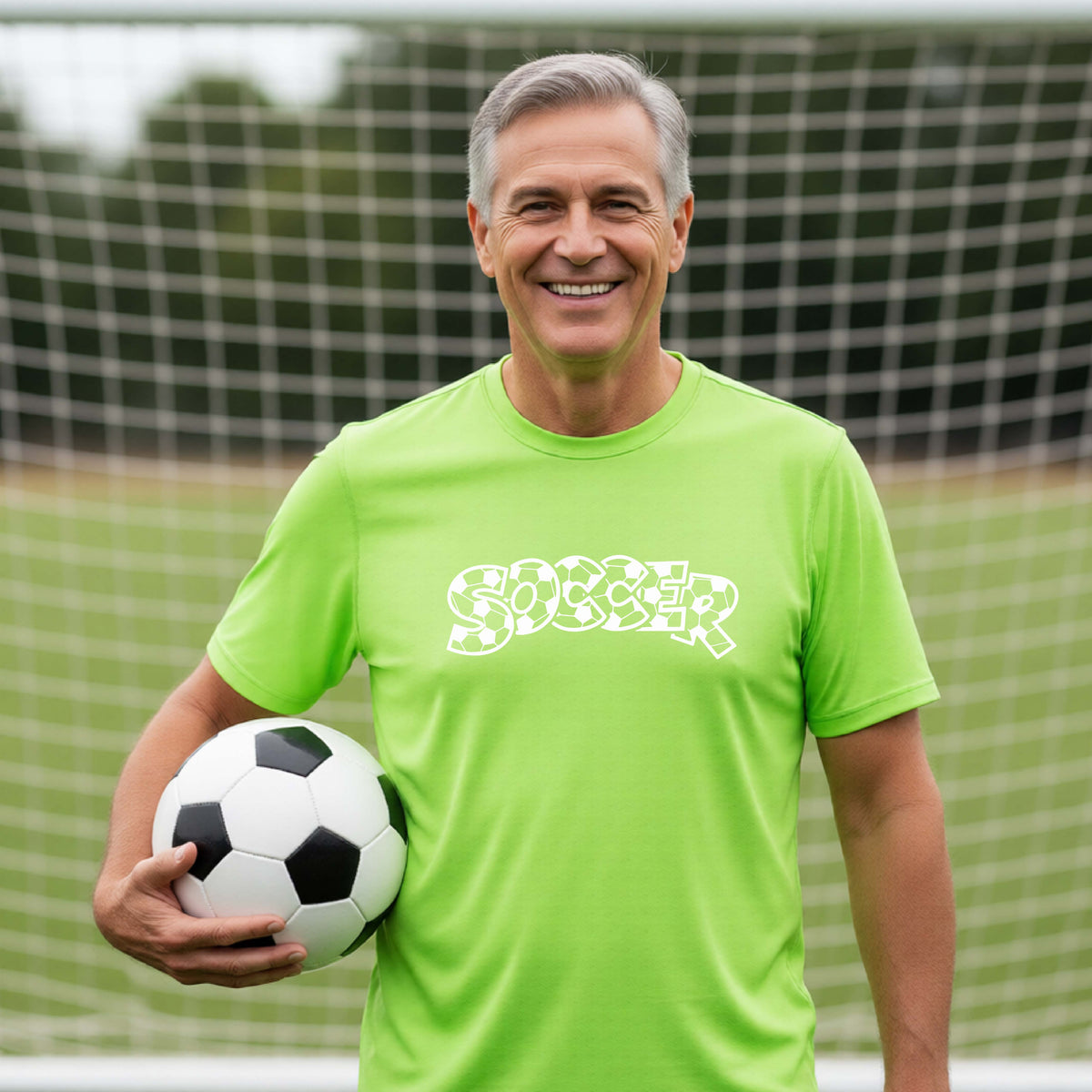 A smiling man wearing a lime green t-shirt, standing on a soccer field in front of a goal net. with a white graphic of the word 'SOCCER' in a stylish soccer pattern printed on the chest, holding a soccer ball.