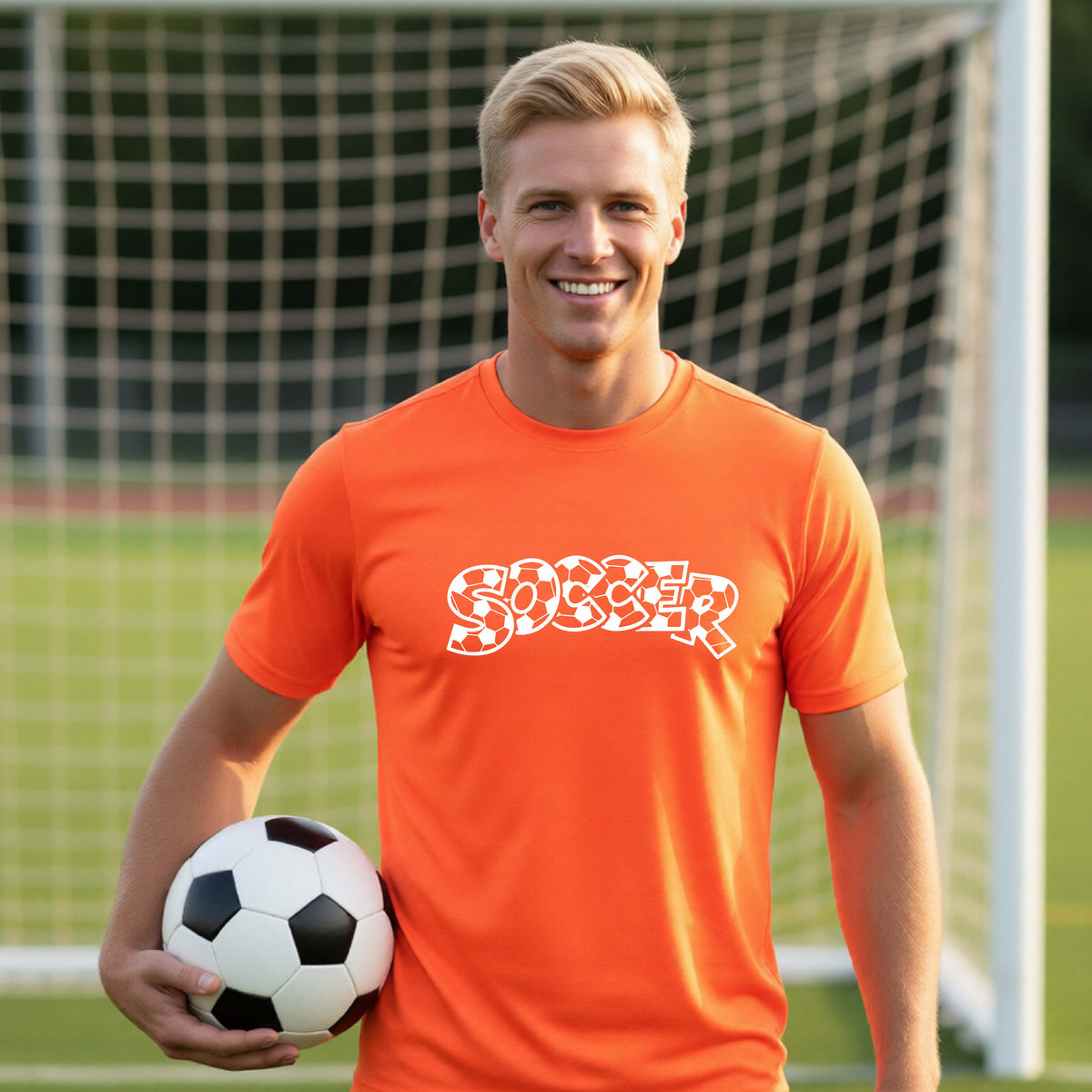 A smiling man wearing an orange t-shirt, standing on a soccer field in front of a goal net. with a white graphic of the word 'SOCCER' in a stylish soccer pattern printed on the chest, holding a soccer ball.