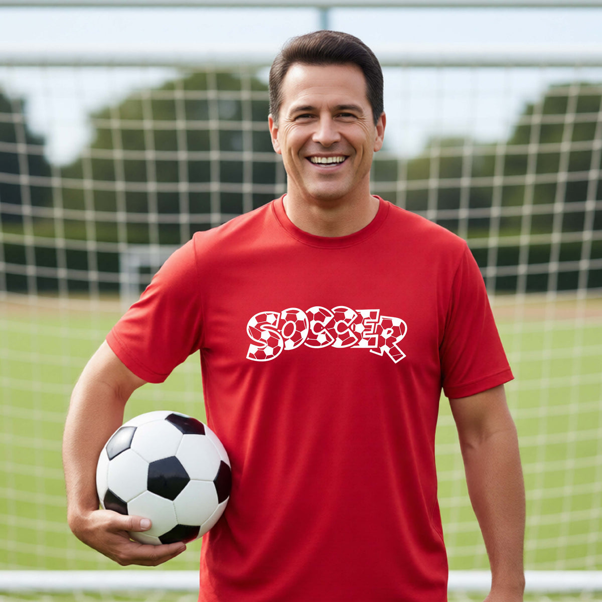 A smiling man wearing a red t-shirt, standing on a soccer field in front of a goal net. with a white graphic of the word 'SOCCER' in a stylish soccer pattern printed on the chest, holding a soccer ball.
