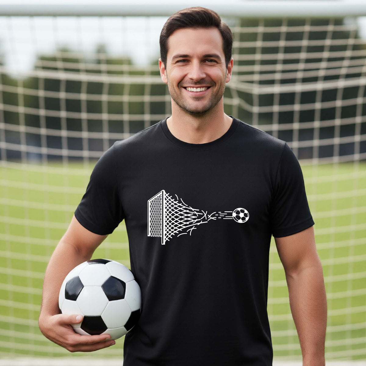 A smiling man wearing a black t-shirt standing on a soccer field in front of a goal net. with a white graphic of a soccer ball shooting through and tearing up a soccer goal net, printed on the chest.