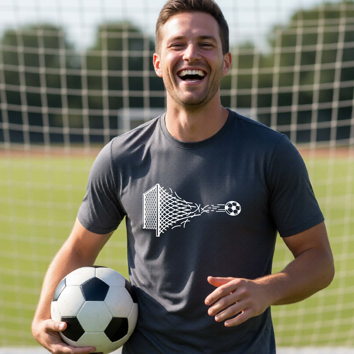 A smiling man wearing a dark gray t-shirt standing on a soccer field in front of a goal net. with a white graphic of a soccer ball shooting through and tearing up a soccer goal net, printed on the chest.