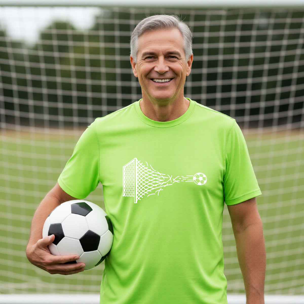 A smiling man wearing a lime green t-shirt standing on a soccer field in front of a goal net. with a white graphic of a soccer ball shooting through and tearing up a soccer goal net, printed on the chest.