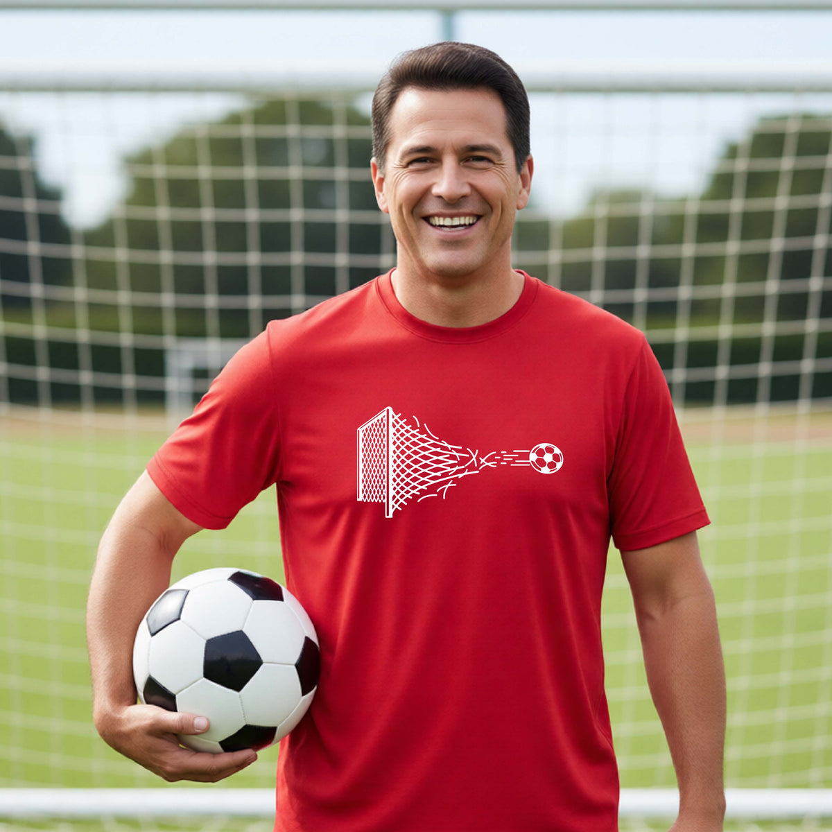 A smiling man wearing a red t-shirt standing on a soccer field in front of a goal net. with a white graphic of a soccer ball shooting through and tearing up a soccer goal net, printed on the chest.