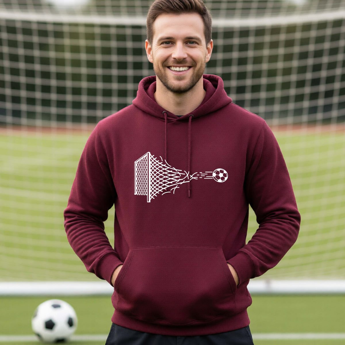 A smiling man wearing a maroon hoodie pullover standing on a soccer field in front of a goal net. with a white graphic of a soccer ball shooting through and tearing up a soccer goal net, printed on the chest.