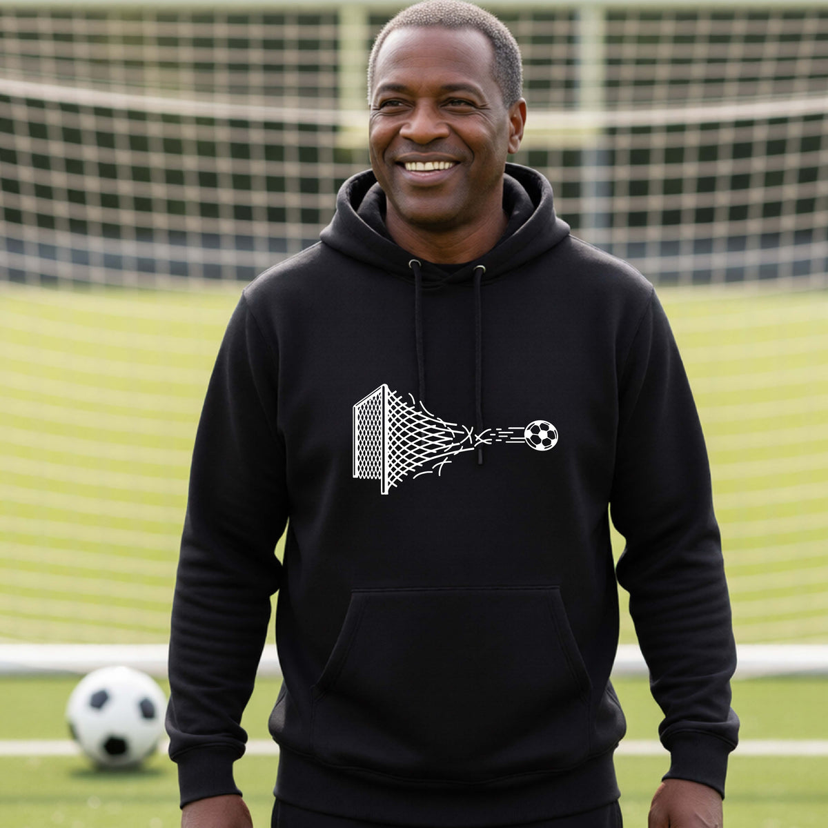 A smiling man wearing a black hoodie pullover standing on a soccer field in front of a goal net. with a white graphic of a soccer ball shooting through and tearing up a soccer goal net, printed on the chest.