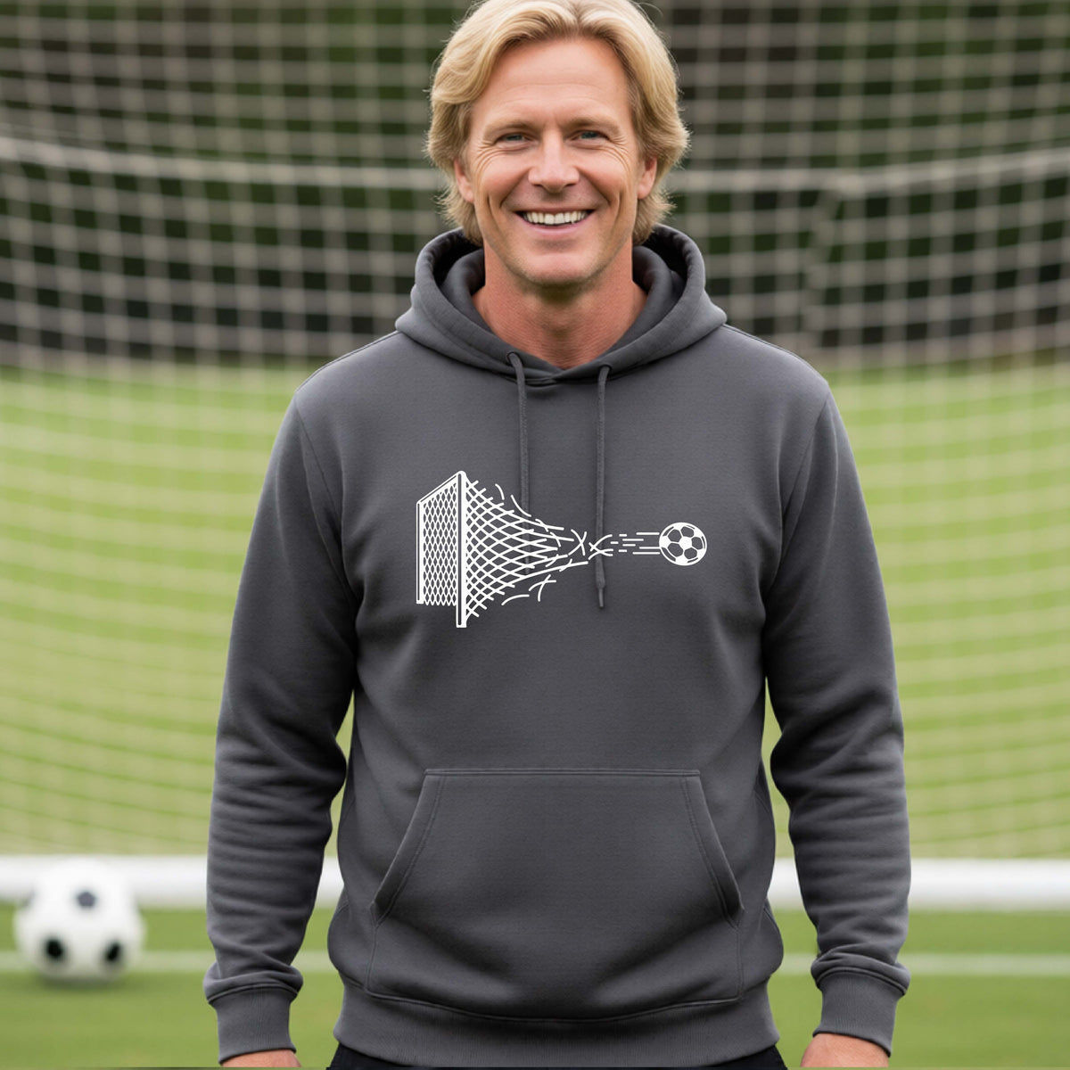 A smiling man wearing a dark gray hoodie pullover standing on a soccer field in front of a goal net. with a white graphic of a soccer ball shooting through and tearing up a soccer goal net, printed on the chest.