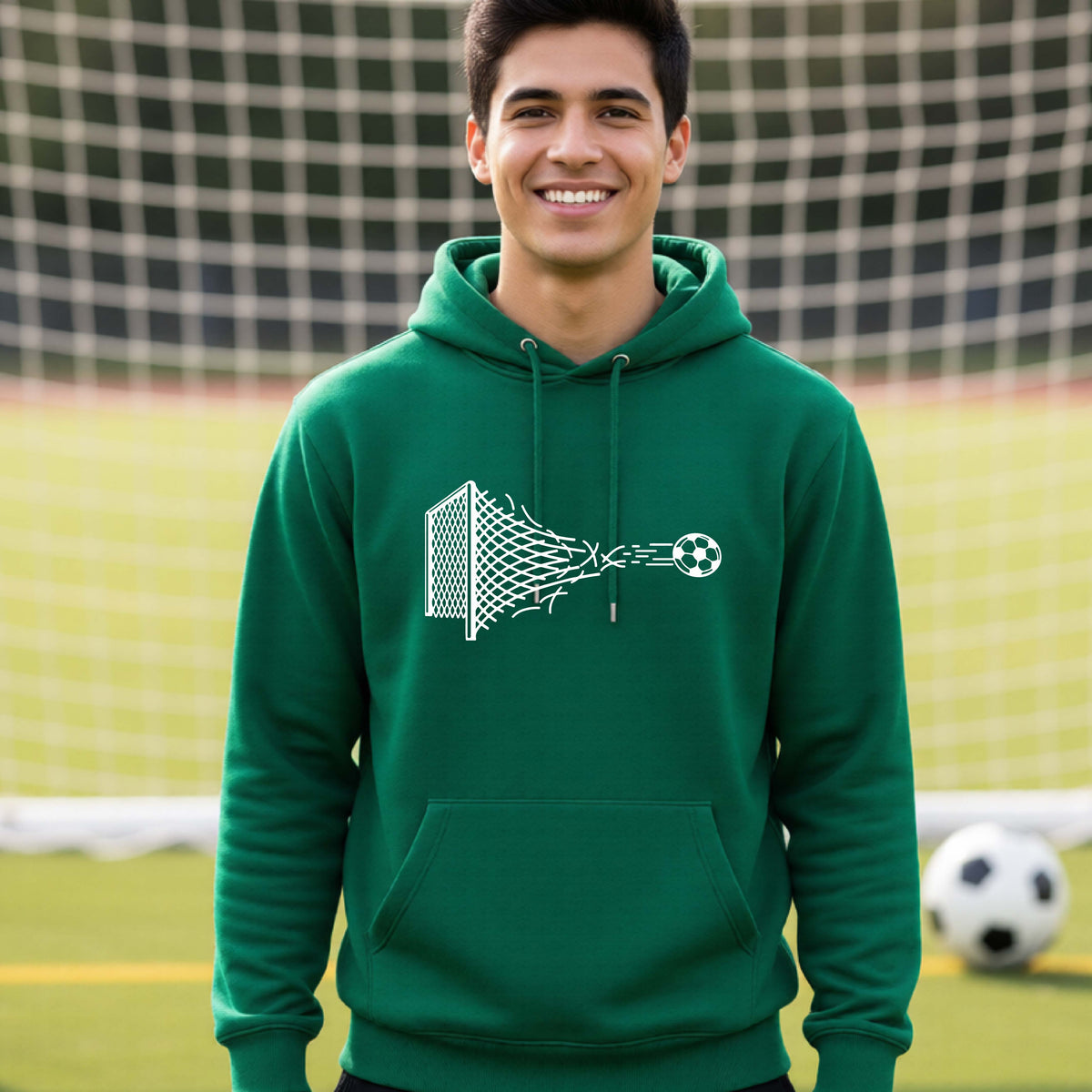 A smiling man wearing a green hoodie pullover standing on a soccer field in front of a goal net. with a white graphic of a soccer ball shooting through and tearing up a soccer goal net, printed on the chest.