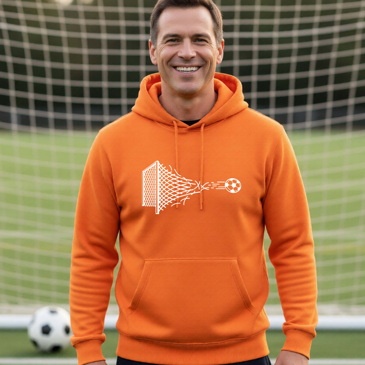 A smiling man wearing an orange hoodie pullover standing on a soccer field in front of a goal net. with a white graphic of a soccer ball shooting through and tearing up a soccer goal net, printed on the chest.