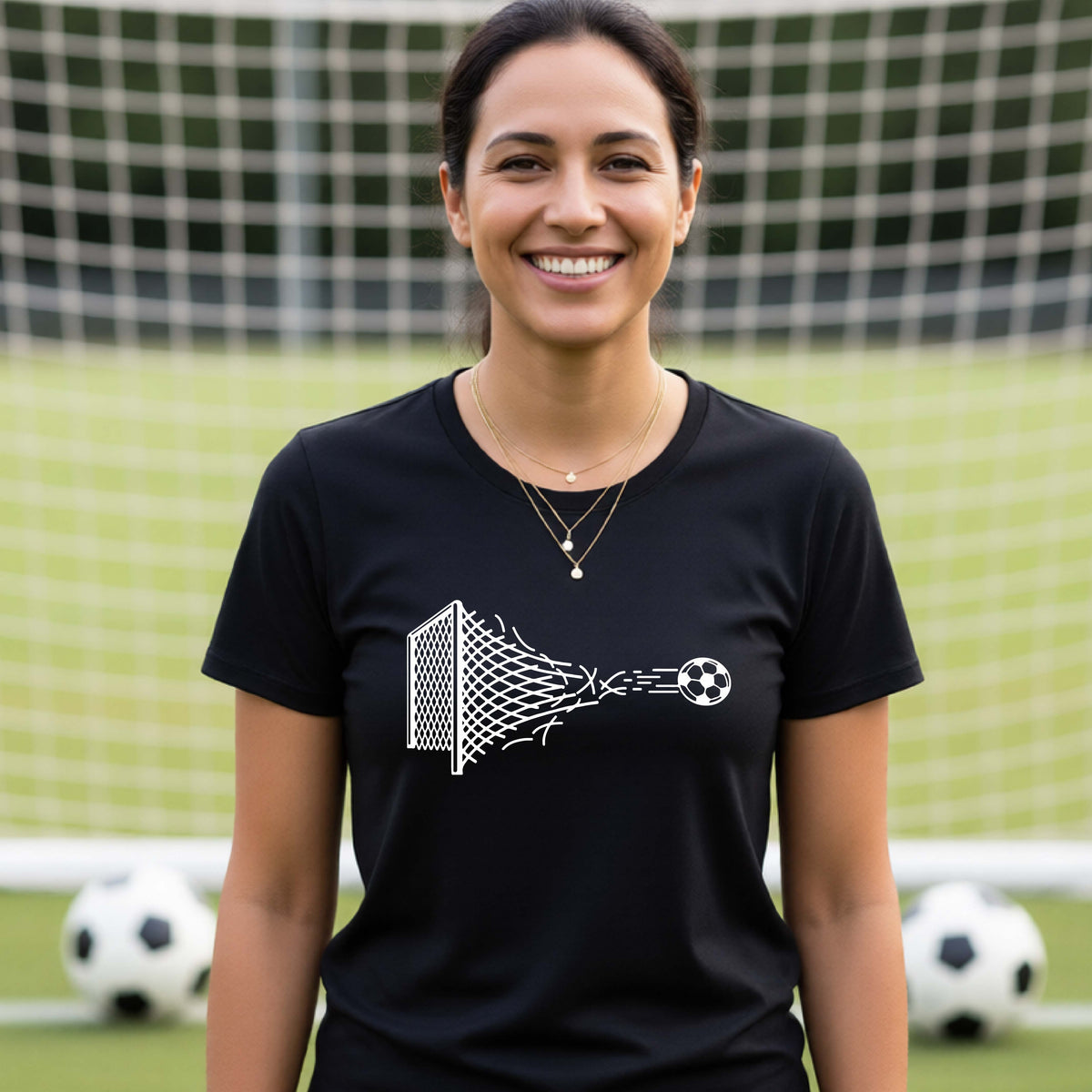 A smiling woman wearing a black t-shirt standing on a soccer field in front of a goal net. with a white graphic of a soccer ball shooting through and tearing up a soccer goal net, printed on the chest.