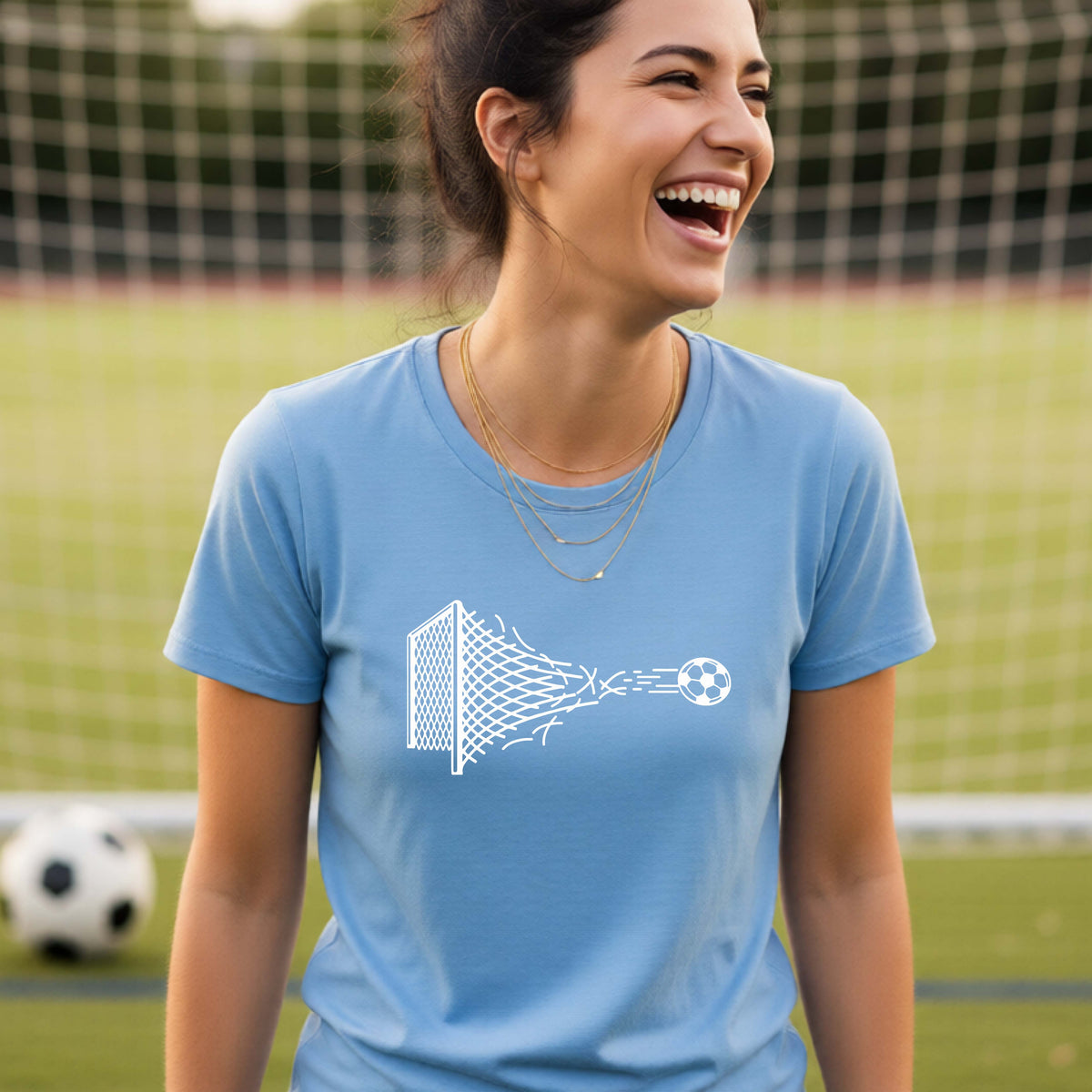 A smiling woman wearing a blue t-shirt standing on a soccer field in front of a goal net. with a white graphic of a soccer ball shooting through and tearing up a soccer goal net, printed on the chest.