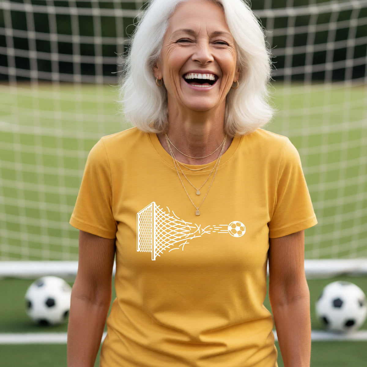 A smiling woman wearing a gold t-shirt standing on a soccer field in front of a goal net. with a white graphic of a soccer ball shooting through and tearing up a soccer goal net, printed on the chest.