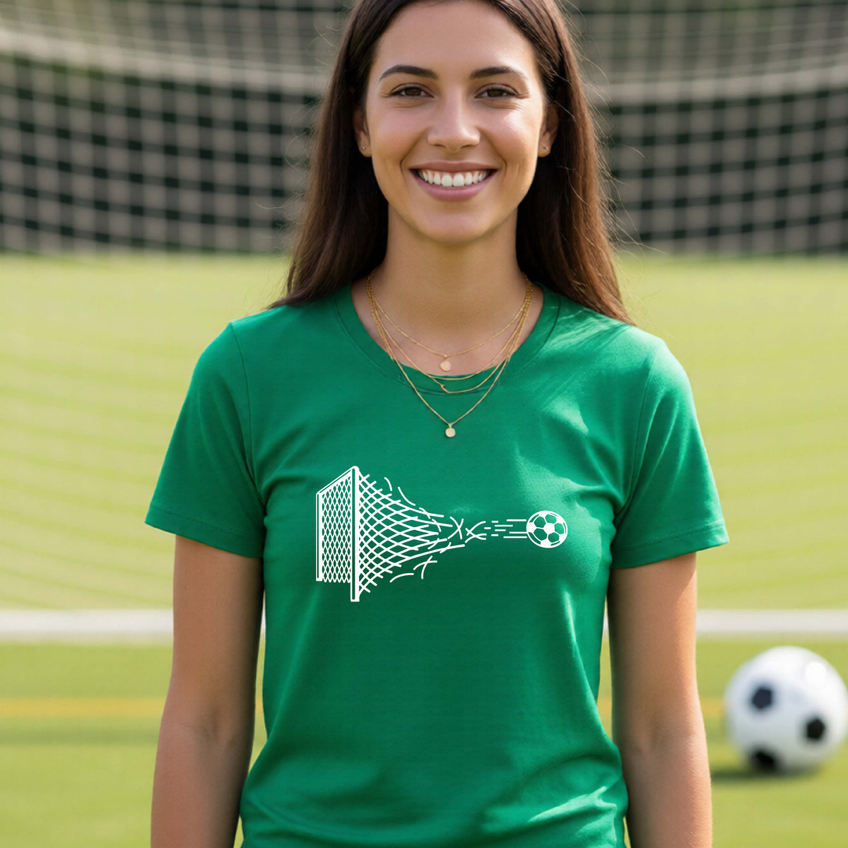 A smiling woman wearing a green t-shirt standing on a soccer field in front of a goal net. with a white graphic of a soccer ball shooting through and tearing up a soccer goal net, printed on the chest.