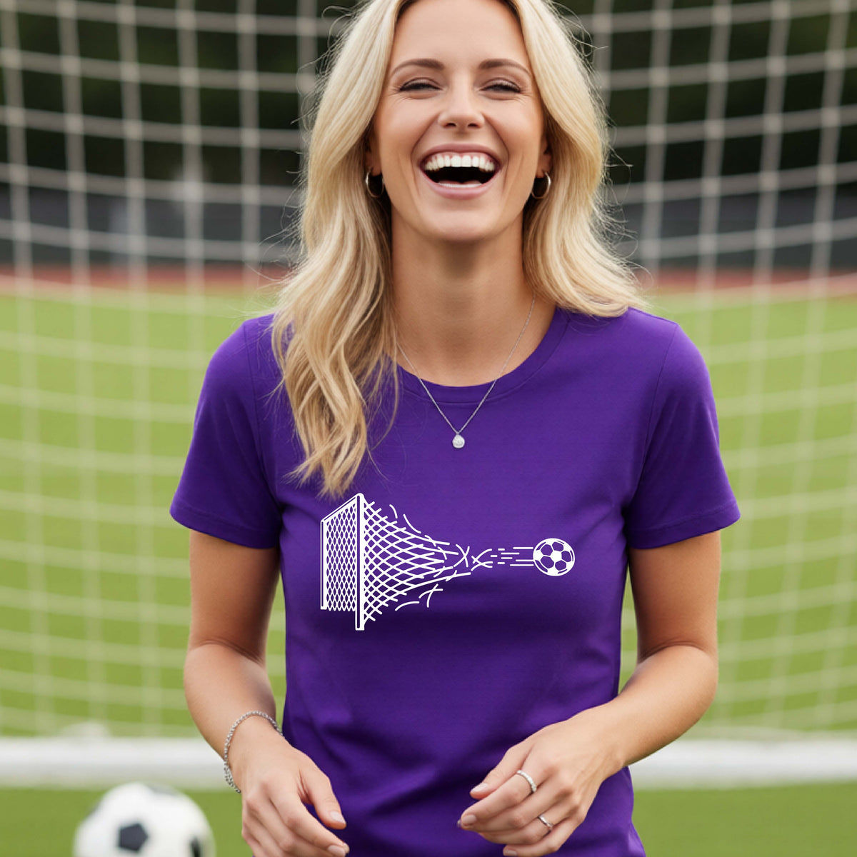 A smiling woman wearing a purple t-shirt standing on a soccer field in front of a goal net. with a white graphic of a soccer ball shooting through and tearing up a soccer goal net, printed on the chest.