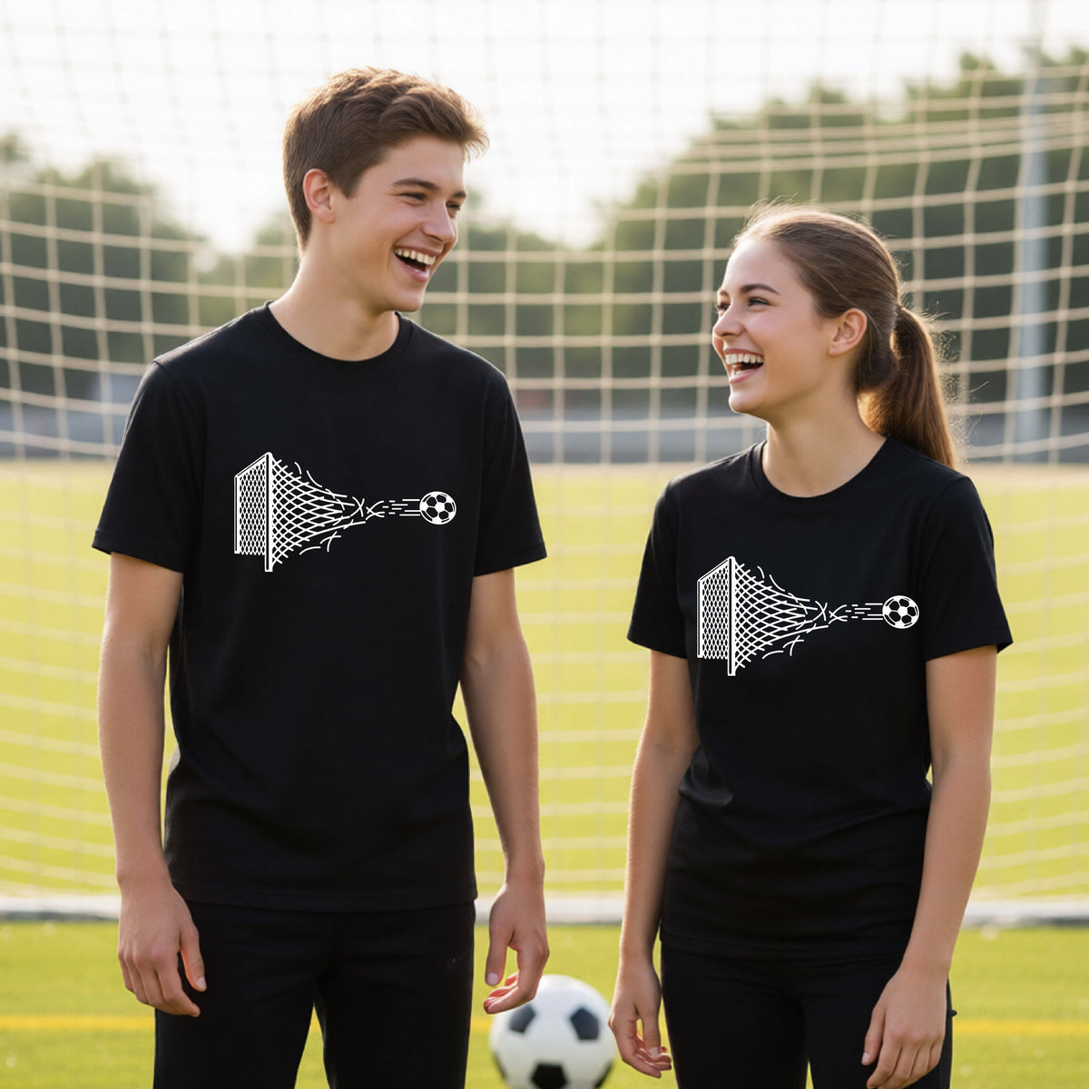 Two smiling young athletes wearing matching black t-shirts standing on a soccer field in front of a goal net. with a white graphic of a soccer ball shooting through and tearing up a soccer goal net, printed on the chest.