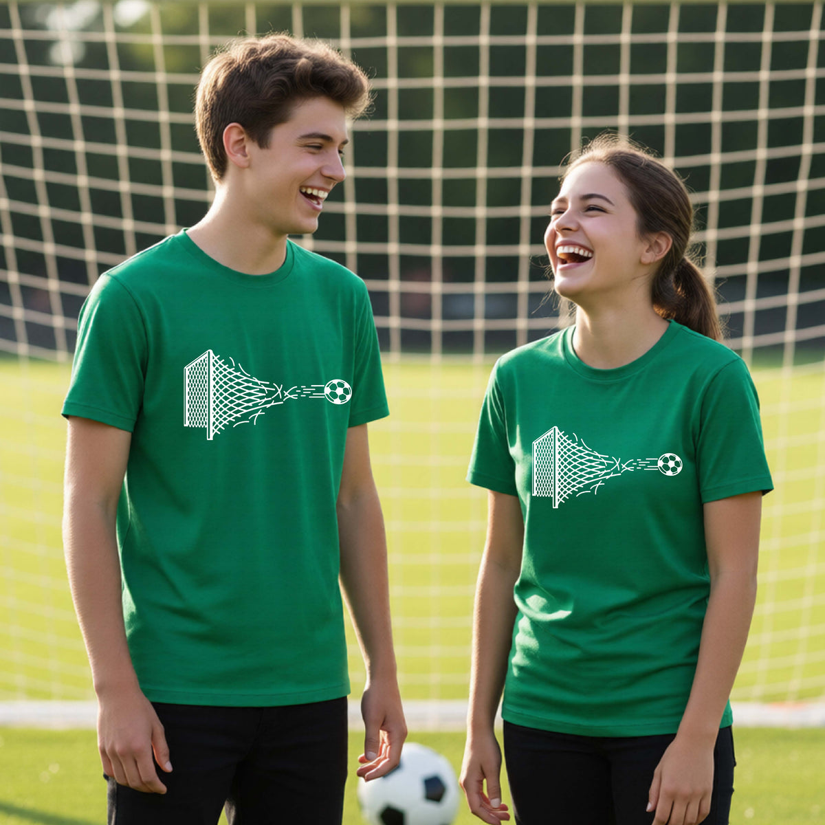Two smiling young athletes wearing matching green t-shirts standing on a soccer field in front of a goal net. with a white graphic of a soccer ball shooting through and tearing up a soccer goal net, printed on the chest.