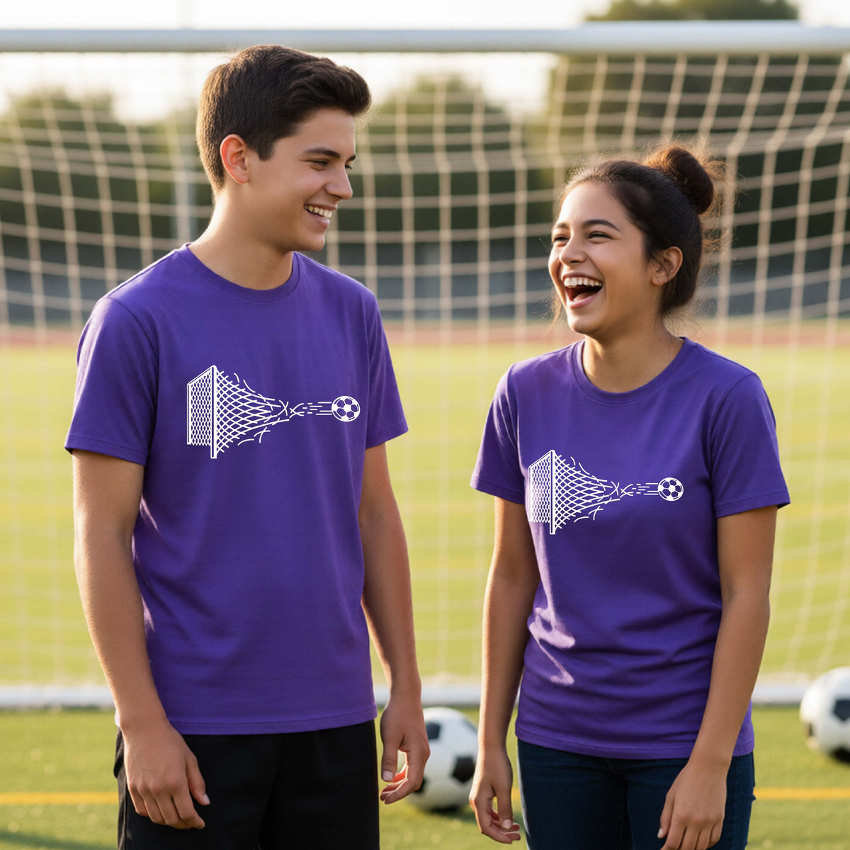 Two smiling young athletes wearing matching purple t-shirts standing on a soccer field in front of a goal net. with a white graphic of a soccer ball shooting through and tearing up a soccer goal net, printed on the chest.