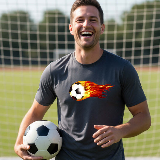 A smiling man wearing a dark gray short-sleeve t-shirt standing on a soccer field in front of a goal net. with a white graphic of a soccer ball shooting through air and trailing yellow, red and orange flames behind it, printed on the chest.