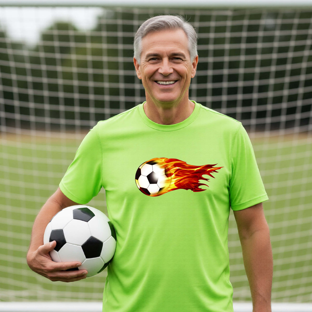 A smiling man wearing a lime green short-sleeve t-shirt standing on a soccer field in front of a goal net. with a white graphic of a soccer ball shooting through air and trailing yellow, red and orange flames behind it, printed on the chest.
